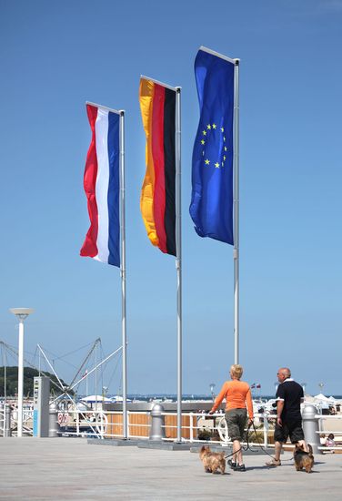 Eine deutsche, niederländische und europäische Fahne wehen an einem Fahnenmast am Strand im Wind