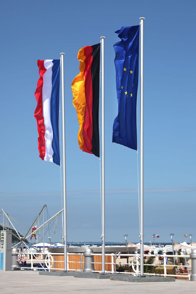 Eine deutsche, niederländische und europäische Fahne wehen an einem Fahnenmast am Strand im Wind