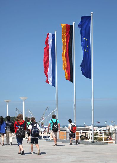 Eine deutsche, niederländische und europäische Fahne wehen an einem Fahnenmast am Strand im Wind