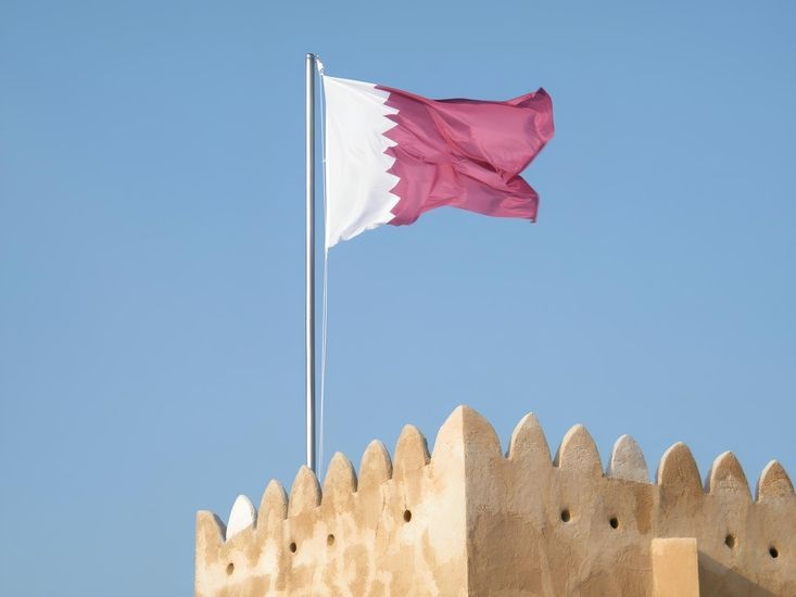 Eine Flagge von Katar an einem Mast auf einem Lehmhaus vor blauem Himmel im Hintergrund