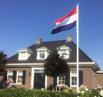 Ein GFK Fahnenmast mit der Flagge von den Niederlanden steht vor einem alten Haus mit blauem Himmel im Hintergrund
