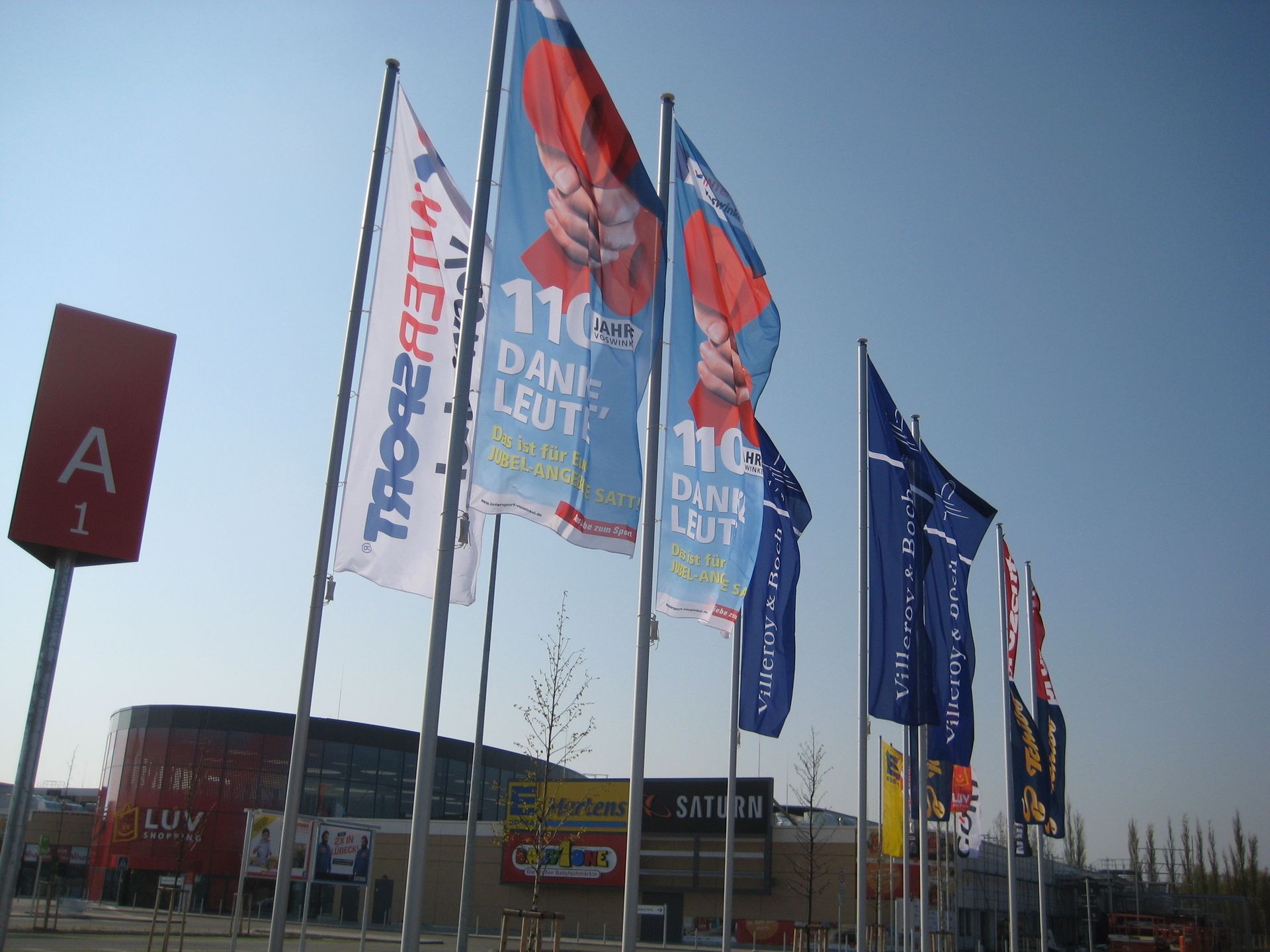 Mehrere Fahnenmäste mit Fahnen wehen auf einem Parkplatz im Wind